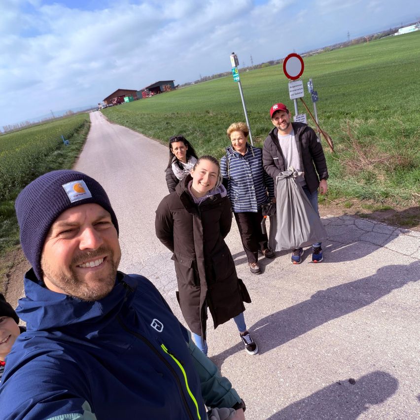 Five people, smiling, stand on a road under a partly cloudy sky. They are surrounded by green fields and some signs.