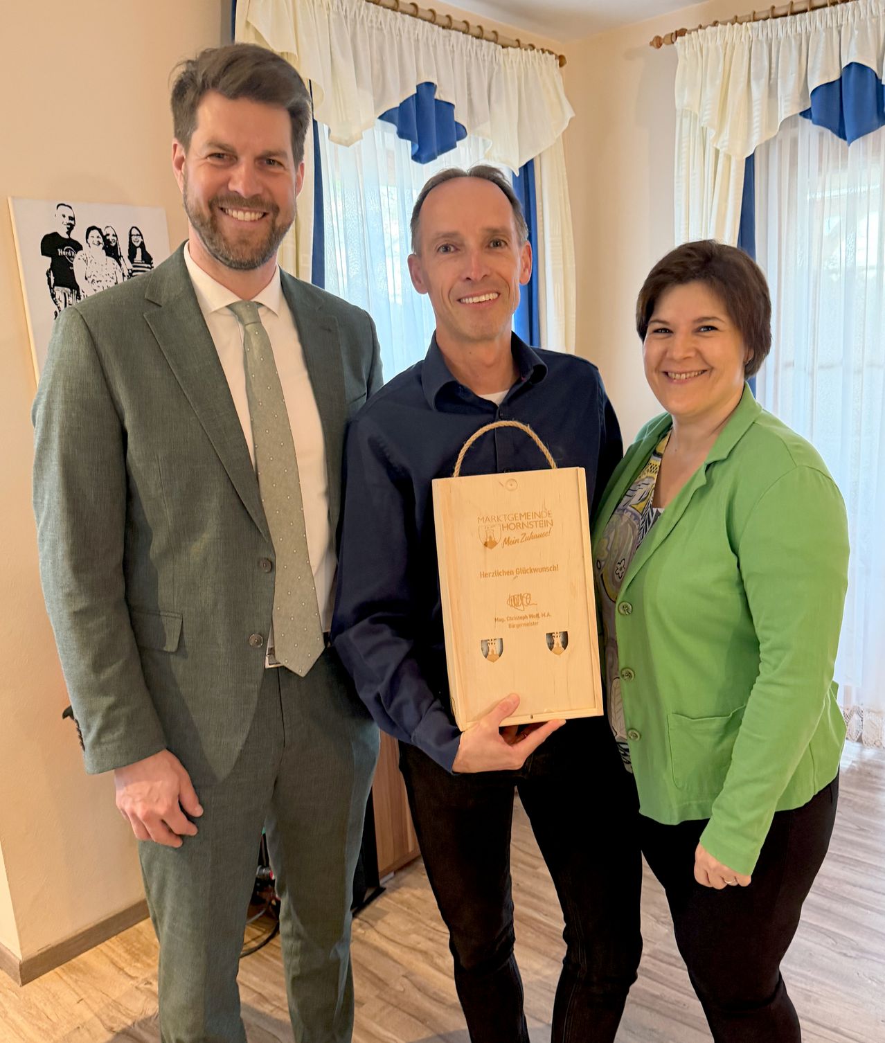 Drei Personen stehen in einem Raum. Zwei Männer und eine Frau. Ein Mann hält eine Holzkiste und scheint sie zu präsentieren. Sie alle lächeln und schauen zur Kamera.