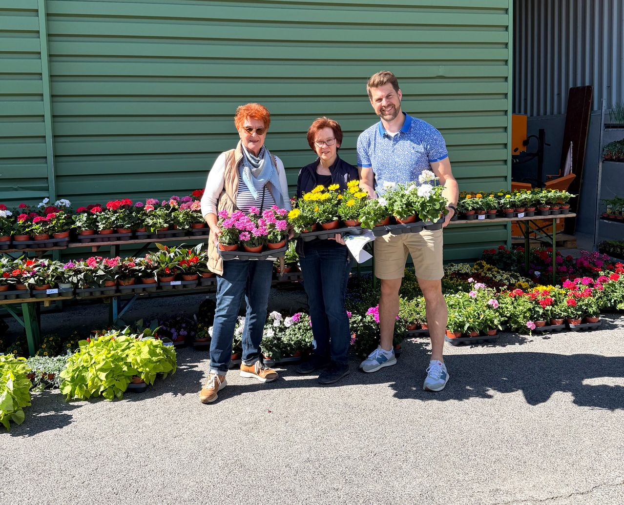 Drei Personen stehen in einem Gartenbereich und halten Blumentrays in der Hand. Dahinter sind viele Topfpflanzen ausgestellt.