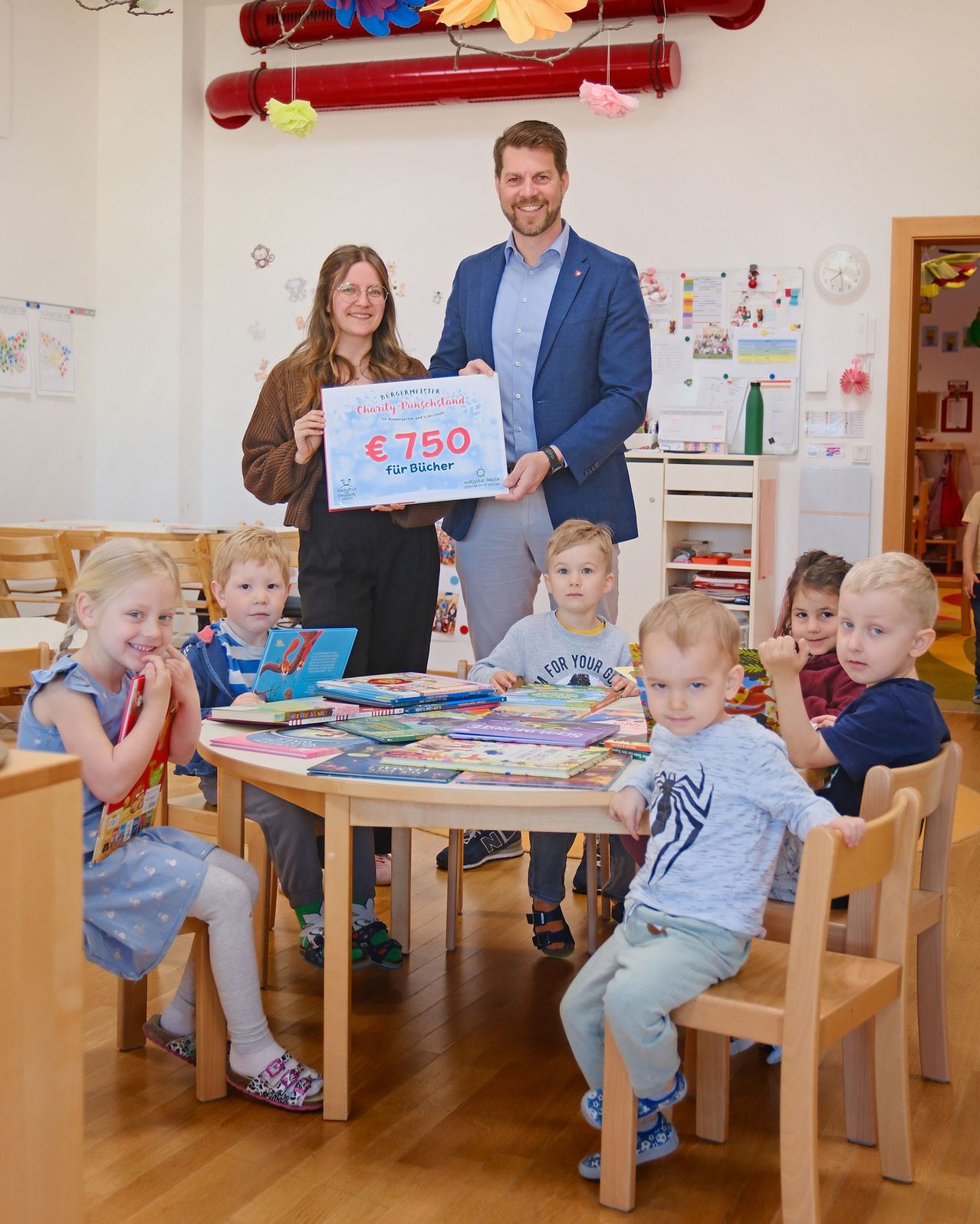 A man and woman stand behind a table of children holding a donation check for 750 euros for books.