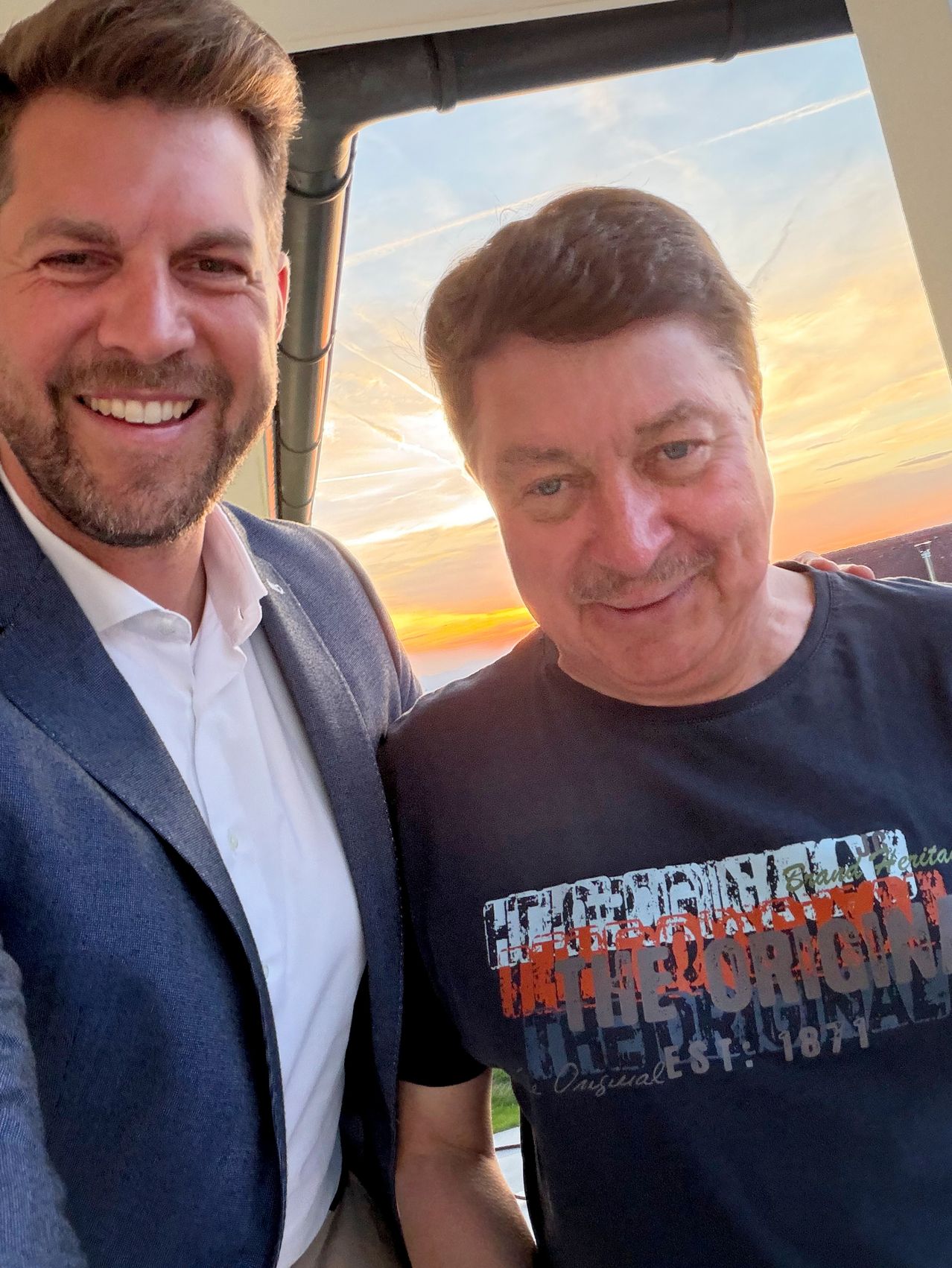 Two men smiling at the camera, one in a suit and the other in a black t-shirt. They are standing outside, with a sunset in the background.