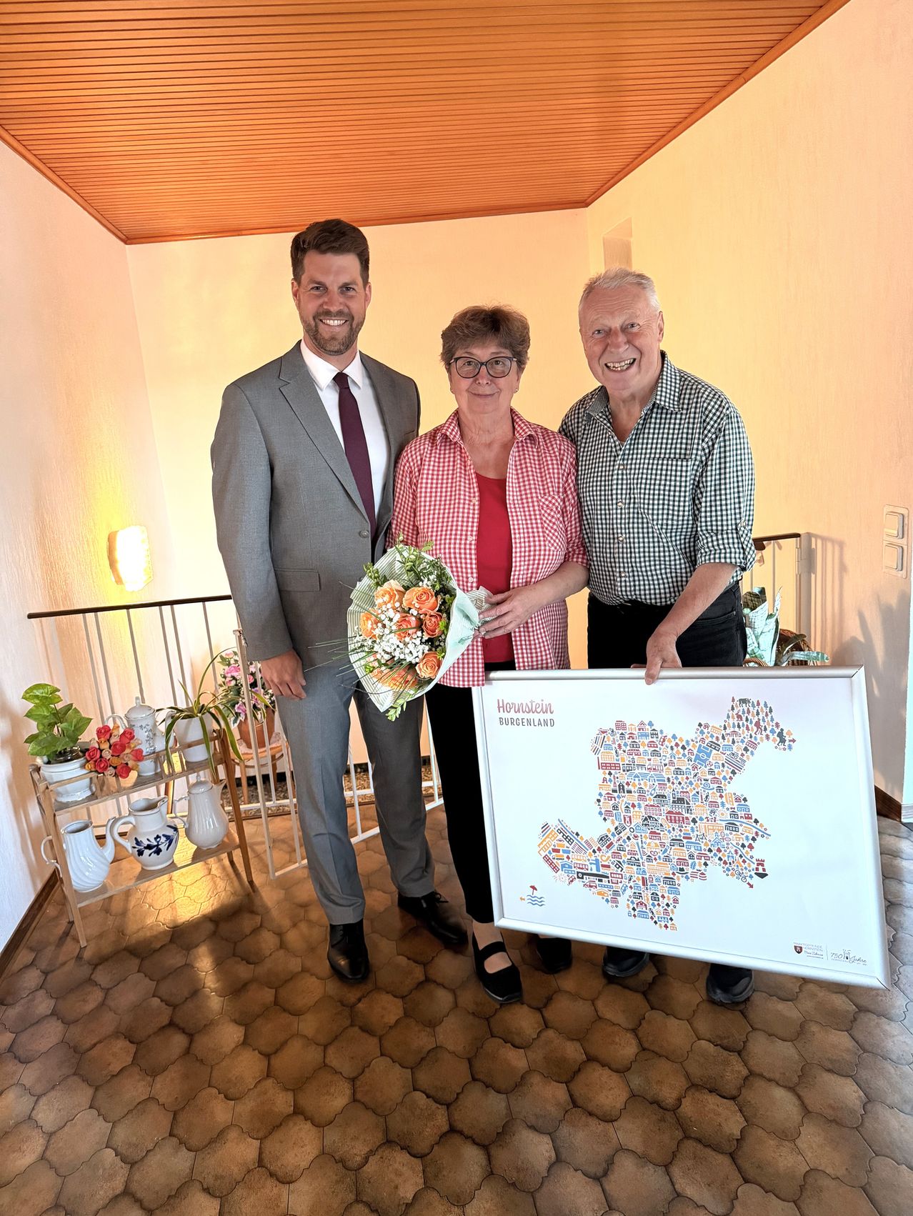 Three people stand in a room holding a framed map of Honstein, Germany. The man on the left wears a gray suit and red tie, the woman in the center holds a bouquet and wears a red checkered shirt, and the man on the right holds the map and wears a checkered shirt.
