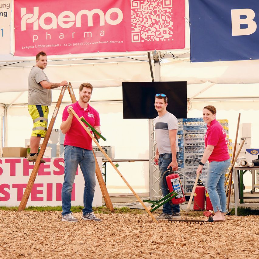 Vier Personen stehen in einem Zelt. Ein Mann klettert eine Leiter hinauf, während ein anderer eine Harke hält. Eine Frau steht neben einem Feuerlöscher. Ein Banner mit dem Text 'Flaemo Pharma' hängt oben.
