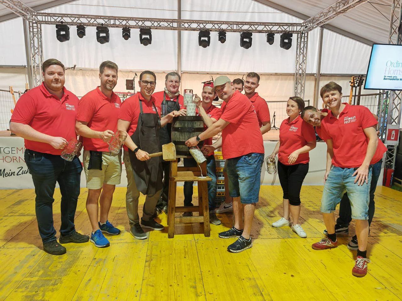 Eine Gruppe von Menschen in roten Shirts, die stehen und lächeln, bereitet sich darauf vor, mit einem Hammer auf einen Fass zu klopfen. Sie stehen auf einem gelben Holzboden, mit einem weißen Zelt und Bühnenbeleuchtung im Hintergrund.