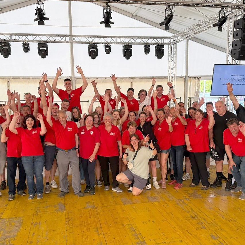 Eine Gruppe von Menschen in roten Hemden und Jeans posiert für ein Foto unter einem Zelt. Einige heben die Hände, lächeln und machen Fotos. Eine große Anzeigetafel ist rechts, und ein Scheinwerfer hängt von der Decke.