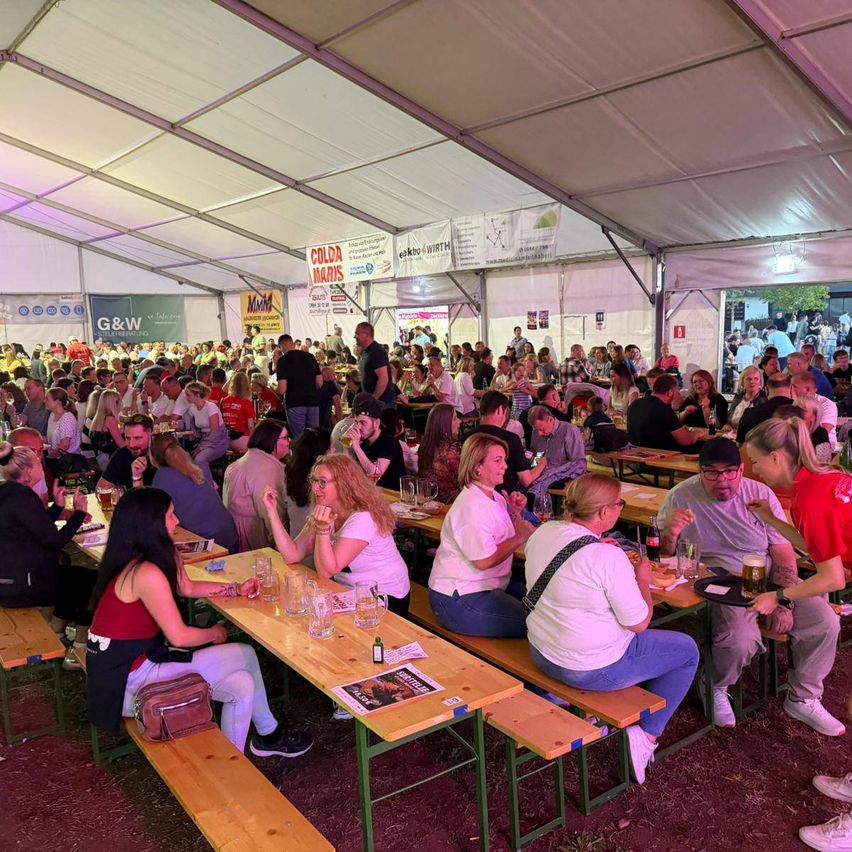 Eine große Menschenmenge versammelt sich in einem Zelt für eine Veranstaltung. Viele sitzen an Tischen und trinken und essen. Einige stehen in der Nähe des Eingangs.