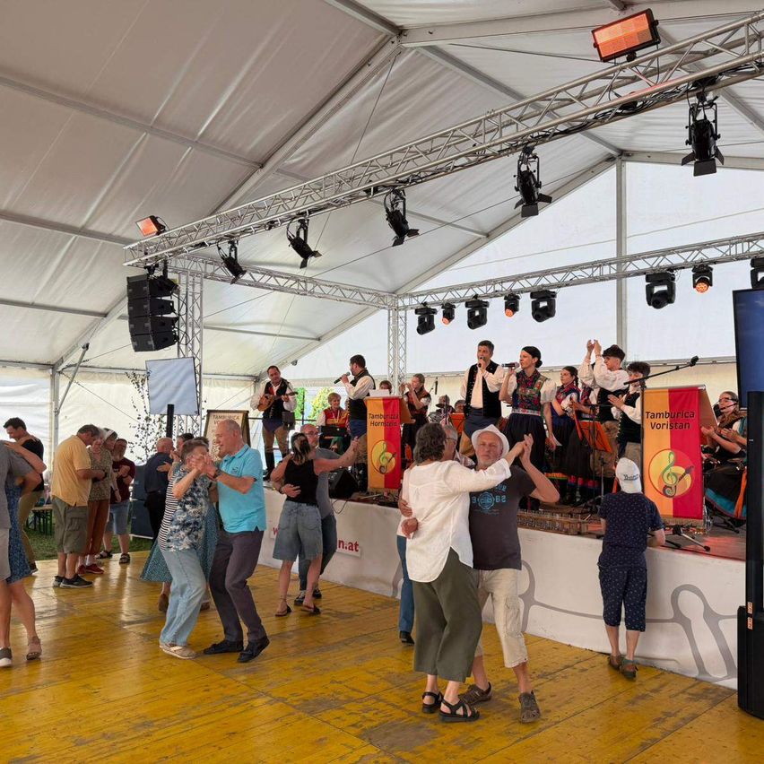 Eine Gruppe von Menschen tanzt in einem großen Zelt mit Bühne und Musikern. Die Bühne ist mit Instrumenten und einem Banner ausgestattet. Es gibt Lichter und Lautsprecher auf der Bühne.