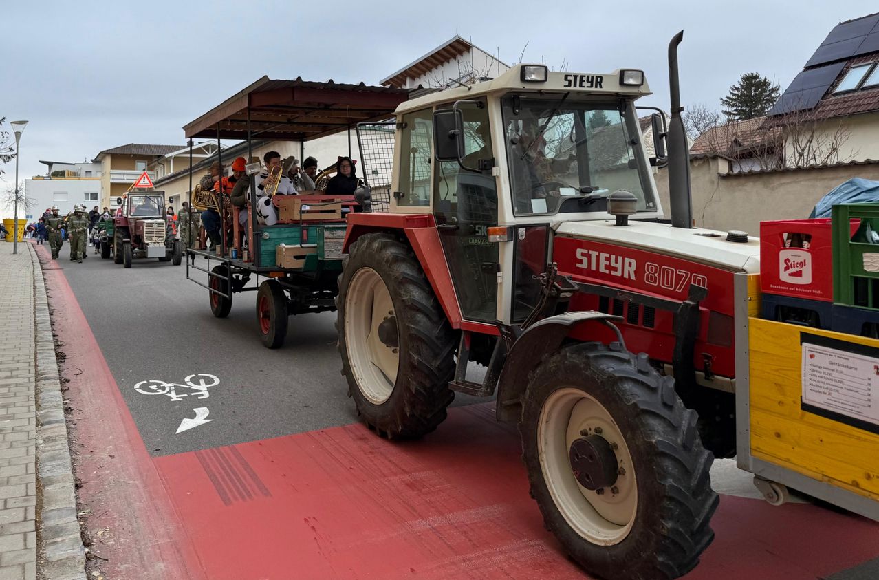 Ein Traktor mit dem Wort Steyr fährt auf der Straße mit einem Anhänger, der mehrere Menschen mit Musikinstrumenten transportiert.