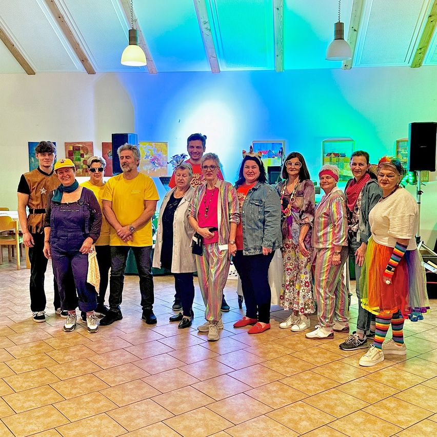 Eine Gruppe von Menschen in bunten Kostümen steht in einem Raum mit gefliesten Böden, weißen Wänden und einer blau-grünen Decke.