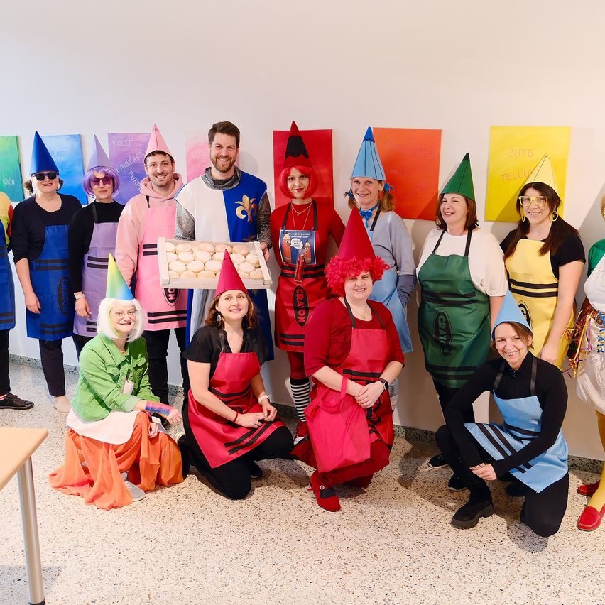 Eine Gruppe von Menschen in bunten Kostümen und Schürzen, möglicherweise für eine thematische Veranstaltung, posiert für ein Foto vor einer weißen Wand mit Plakaten. Einige von ihnen halten eine Schachtel mit Essen.