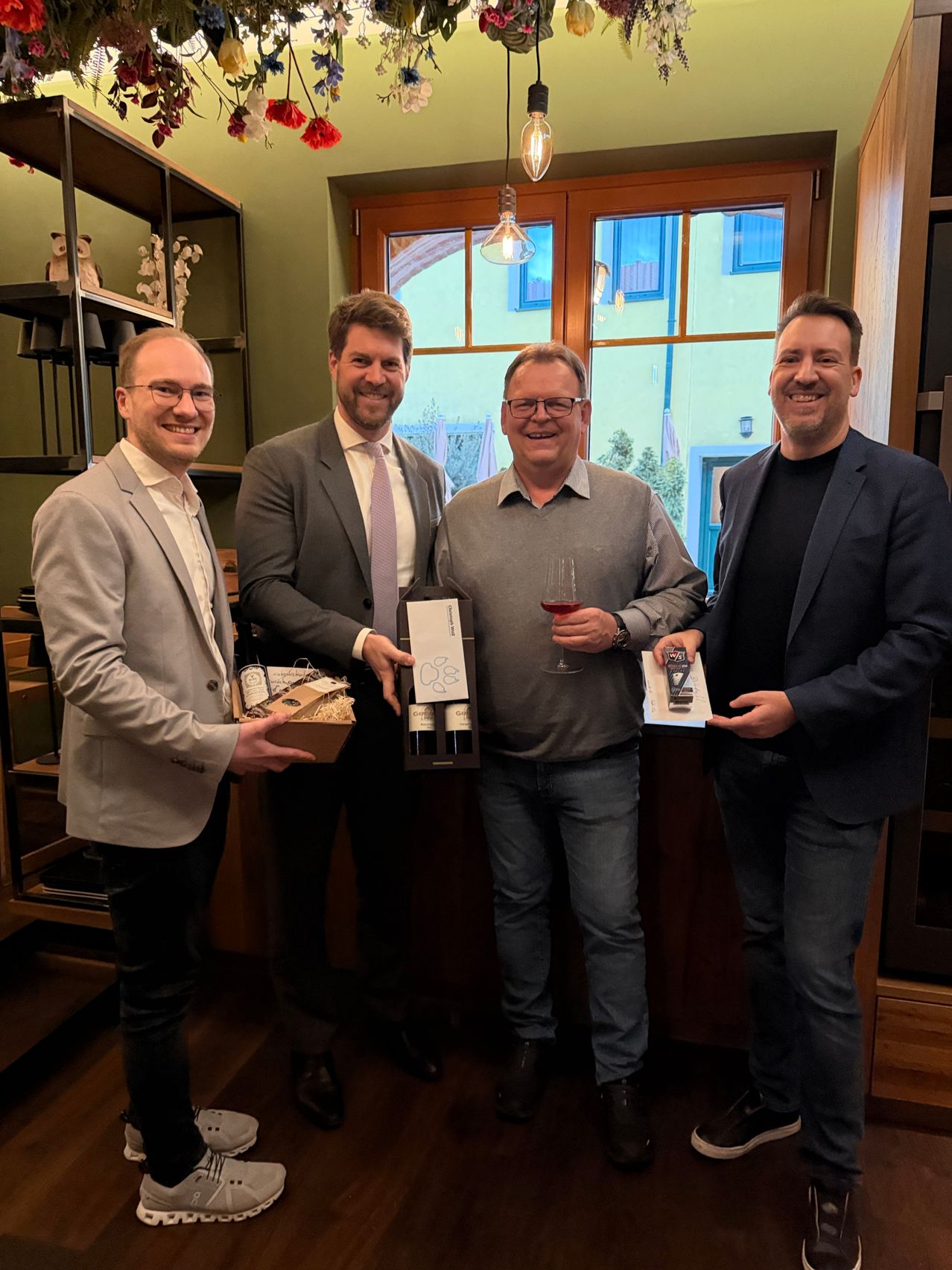 Vier Männer stehen in einem Raum, halten Weinflaschen und Schachteln und lächeln für ein Foto. Der Mann in der Mitte hält ein Weinglas.