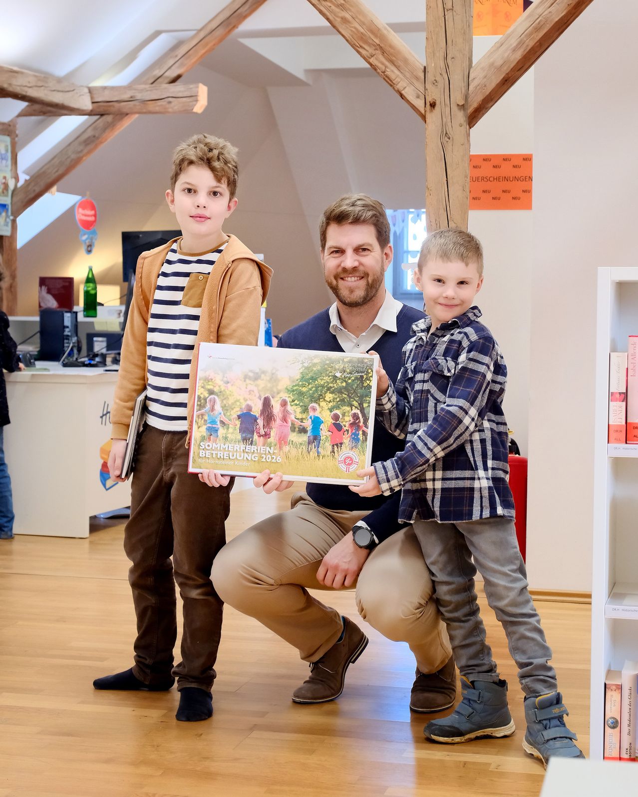 Ein Mann kniet sich hin und hält ein Buch mit zwei Jungen, die in einem Raum mit Bücherregalen, einem Schreibtisch und einem Schild lächelnd in die Kamera schauen.