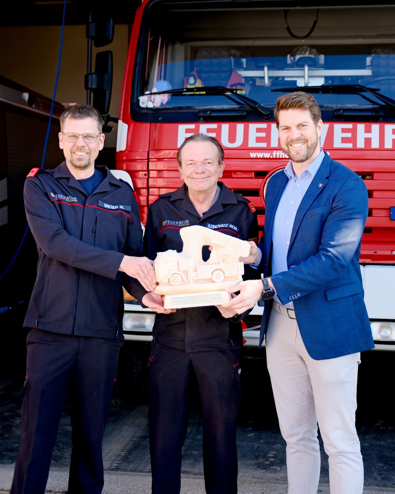 Drei Männer stehen lächelnd vor einem roten Feuerwehrauto und halten ein hölzerne Modell eines Feuerwehrautos in der Hand. Der Mann auf der linken Seite trägt eine Brille und eine Uniform mit dem Wort 'BRESEN'. Der Mann in der Mitte hat einen Abzeichen auf der Brust. Der Mann auf der rechten Seite trägt einen blauen Anzug und eine Uhr.
