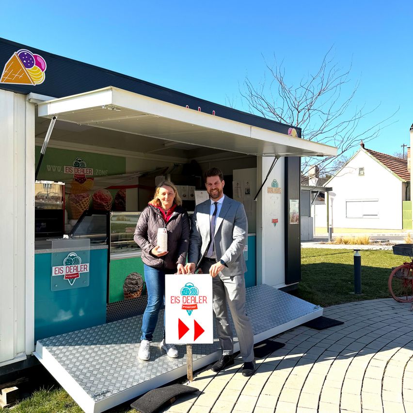 Ein Mann und eine Frau stehen vor einem Eiswagen mit einem Schild, auf dem EIS DEALER steht. Der Wagen ist geöffnet, und sie halten Eisbecher in den Händen.