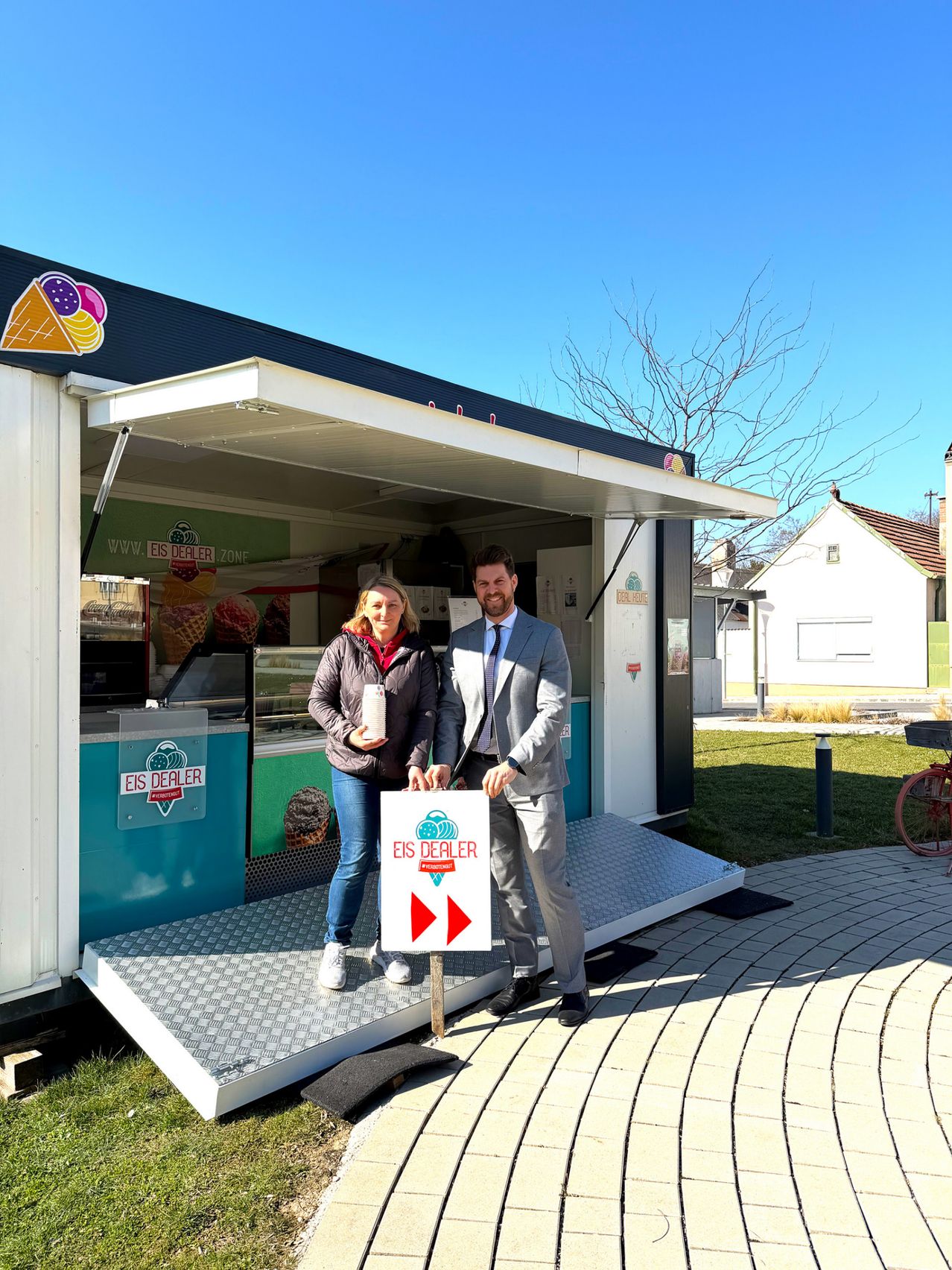 A man and a woman stand outside an ice cream truck with a sign that says EIS DEALER. The truck is open, and they are holding cups of ice cream.