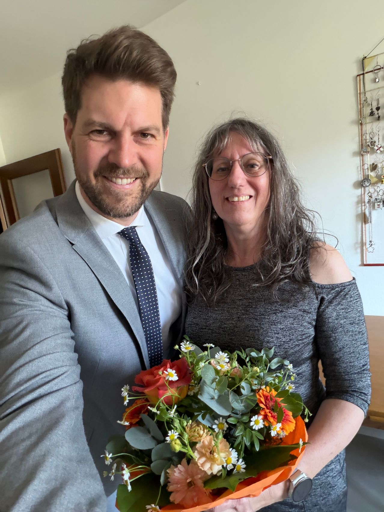 Ein Mann und eine Frau stehen lächelnd nebeneinander. Der Mann ist in einem Anzug und die Frau in einem grauen Kleid. Sie halten einen Blumenstrauß.