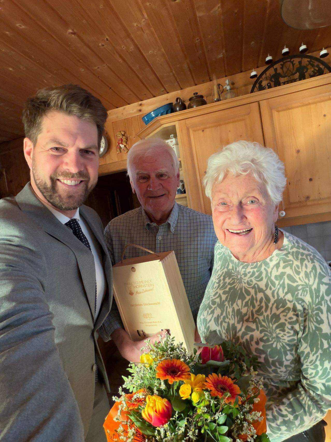 Drei Menschen stehen lächelnd in einer Küche. Ein Mann links hält einen Blumenstrauß, ein anderer Mann eine Schachtel und eine ältere Frau lächelt. Im Hintergrund befinden sich Holzschränke und Regale mit verschiedenen Gegenständen.