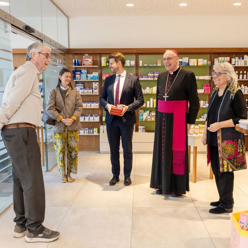 Eine Gruppe von Menschen steht in einer Apotheke. Ein Mann im Anzug hält ein Buch. Ein älterer Mann mit Brille lächelt. Eine Frau in einer Blumenjacke steht hinter ihm.
