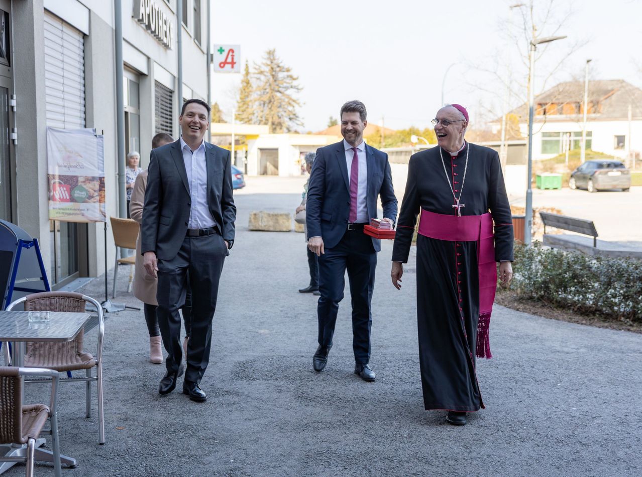 Drei Männer, in formeller Kleidung gekleidet, gehen gemeinsam auf einer Straße. Sie lächeln und scheinen guter Laune zu sein.