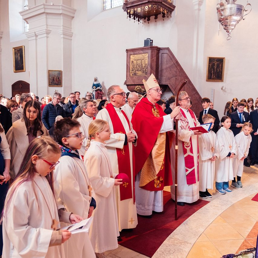 Eine religiöse Zeremonie in einer Kirche, mit mehreren Menschen in weißen und roten Roben. Der Priester hält ein Buch und einen Stab, und es gibt Kinder in der vordersten Reihe.