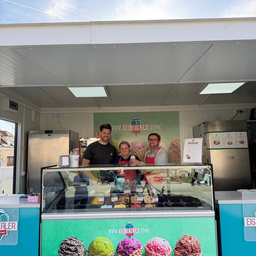 Drei Personen stehen hinter einer Glasvitrine in einem Eisladen, mit verschiedenen Geschmacksrichtungen sichtbar. Ein Schild im Hintergrund lautet 'www.eisdealer.zone'.