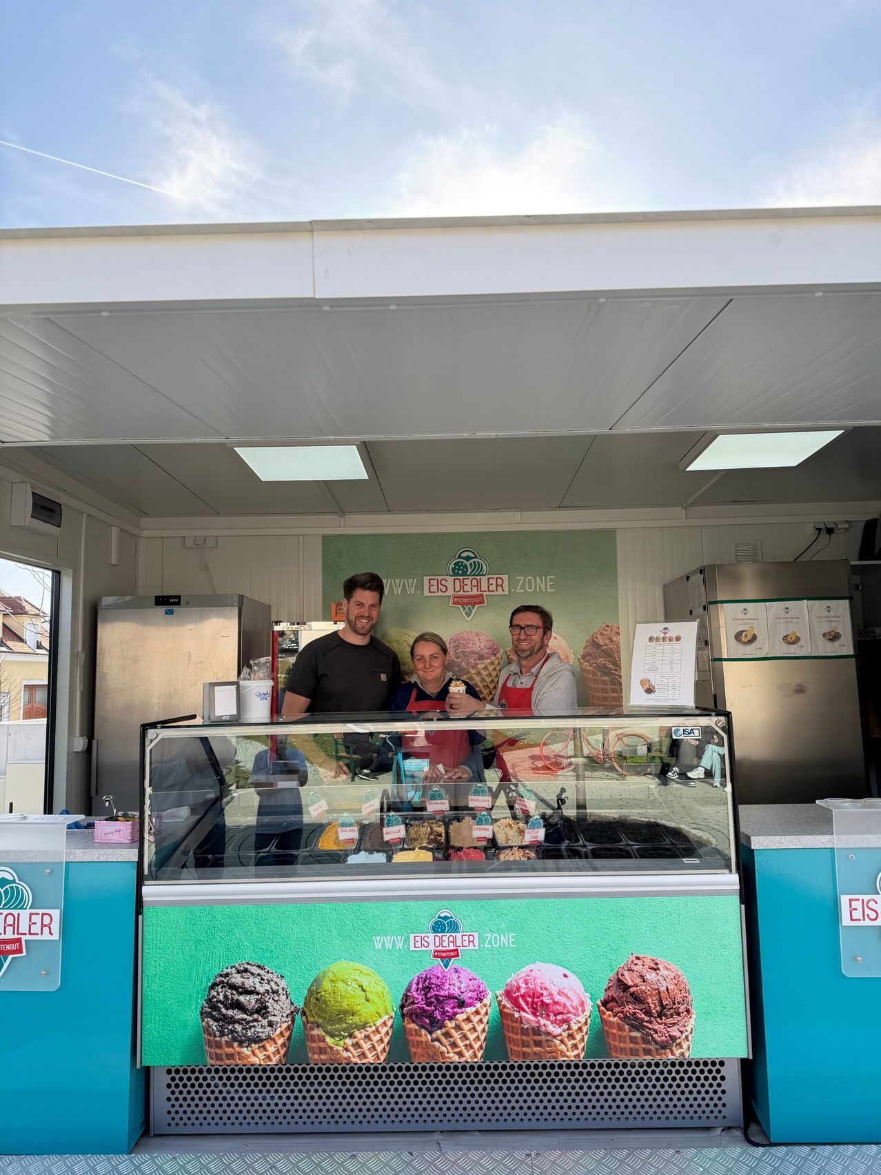 Drei Personen stehen hinter einer Theke in einer Eisdiele. Ein Mann in einem schwarzen T-Shirt lächelt, während eine Frau in einer roten Schürze einen Eisbecher hält. Dahinter zeigt ein großes Poster das Logo und den Namen der Eisdiele.