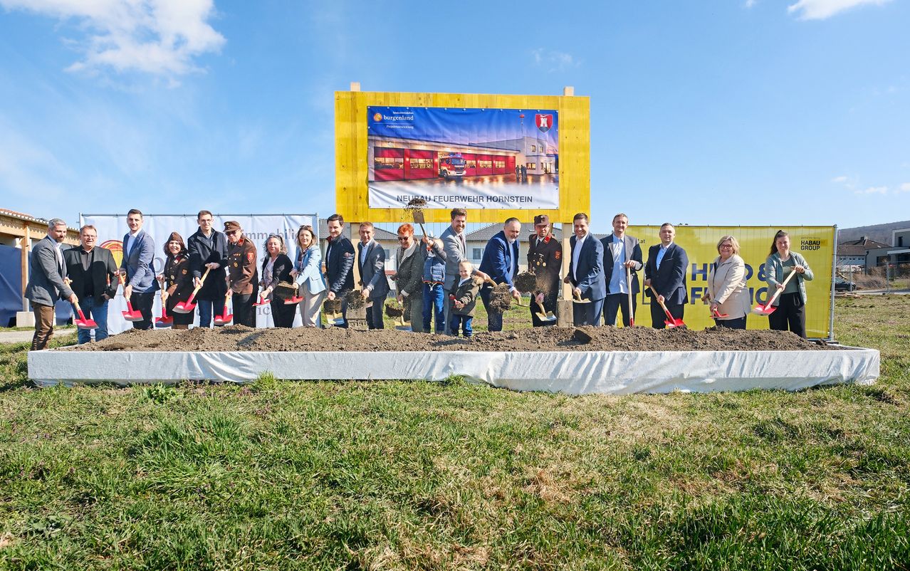 Gruppe von Menschen bei einer Grundsteinlegungszeremonie, die Schaufeln halten und vor einem großen Schild stehen. Dahinter ist ein Banner mit einem Bild einer Feuerwache.