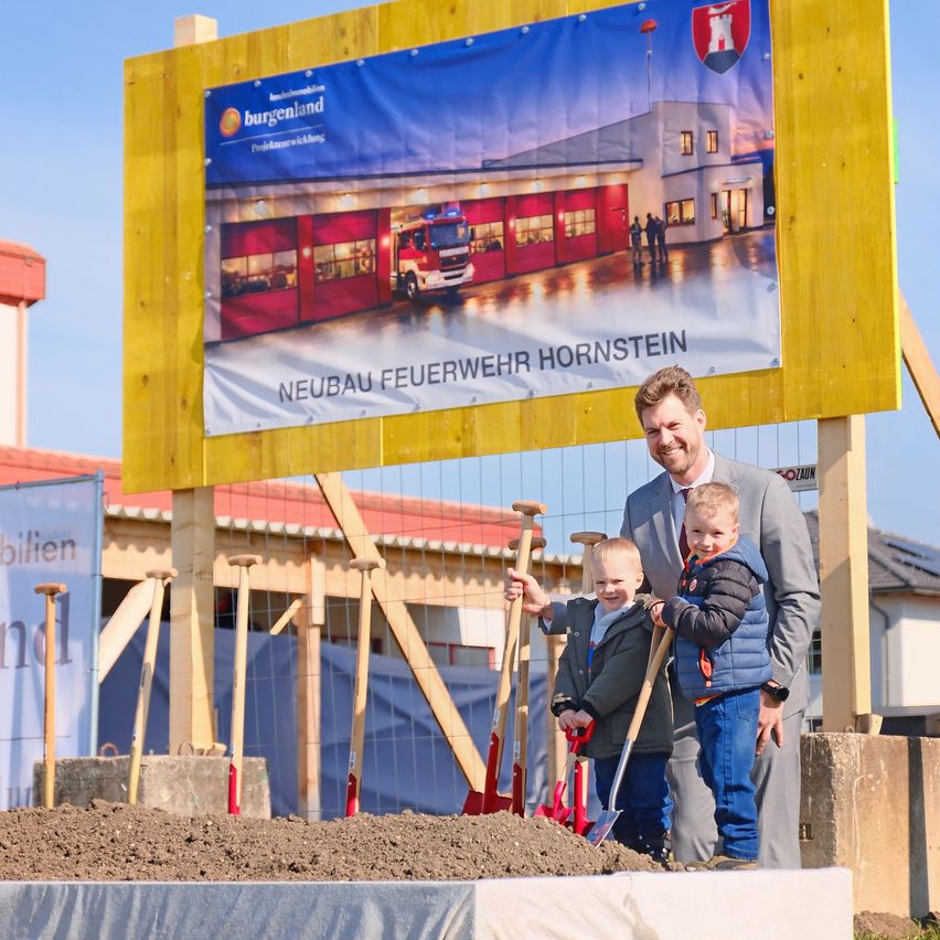 Ein Mann mit zwei kleinen Jungen vor einer Baustelle. Hinter ihnen ein großes Banner mit den Worten 'Neubau Feuerwehr Hornstein'. Sie halten Schaufeln in den Händen.
