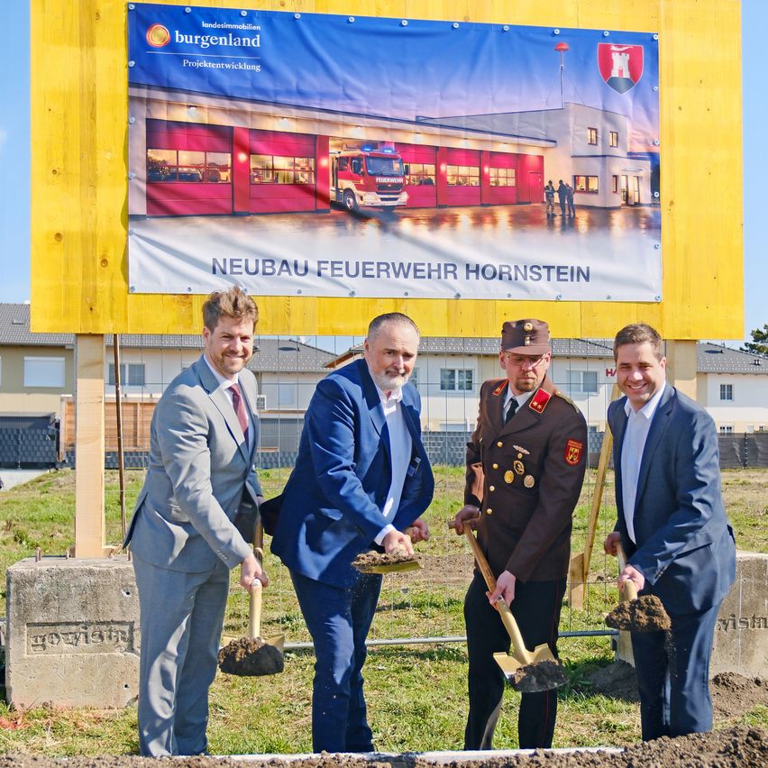 Vier Männer in Anzügen halten Schaufeln vor einem gelben Schild, auf dem 'Neubau Feuerwehr Hornstein' steht. Dahinter befindet sich eine große Feuerwache.