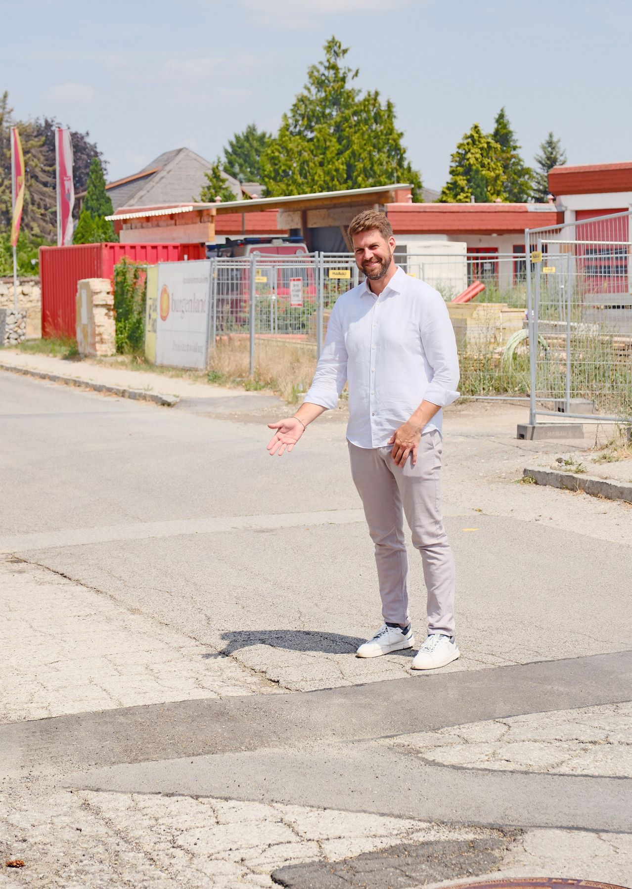 Ein Mann in einem weißen Hemd und grauen Hosen steht auf einer Straße und gestikuliert mit den Händen. Hinter ihm befinden sich Bauzäune, ein rotes Gebäude und ein Fahnenmast.