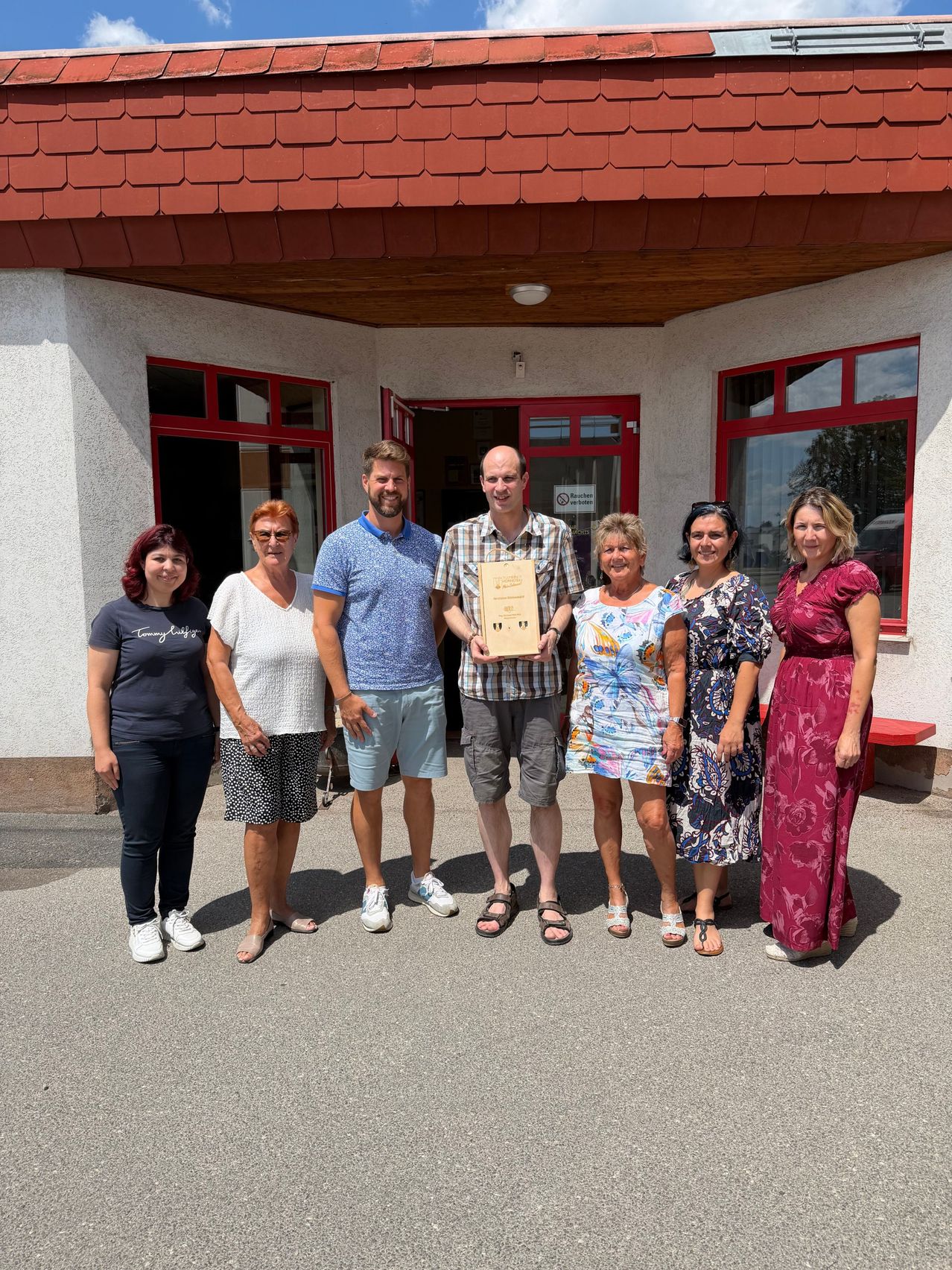 Eine Gruppe von Menschen steht vor einem Gebäude, zwei Männer halten eine Urkunde und posieren für ein Foto.