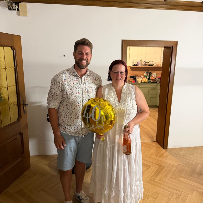 Ein Mann und eine Frau stehen in einem Zimmer, lächeln und halten einen goldenen Luftballon mit der Aufschrift 40. Geburtstag. Die Frau hält eine Flasche Wein. Hinter ihnen befindet sich eine Küche mit einer Holztür.