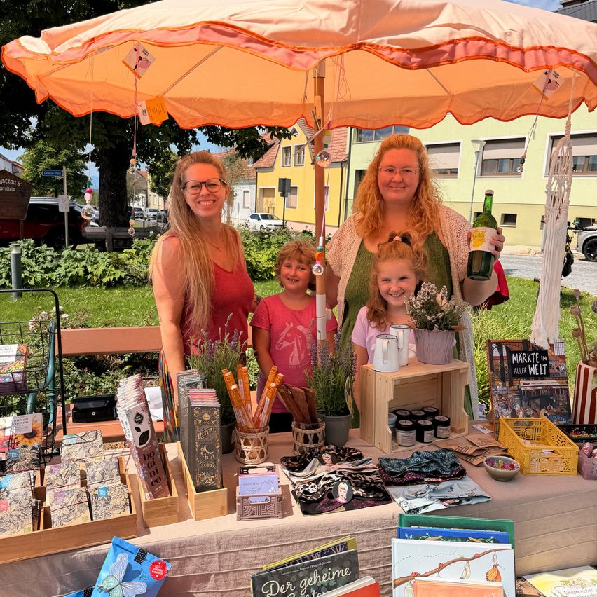 Drei Frauen und ein junges Mädchen stehen unter einem orangefarbenen Regenschirm an einem Marktstand und lächeln. Der Stand ist mit verschiedenen Artikeln wie Büchern, Blumen und Handwerksarbeiten gefüllt.