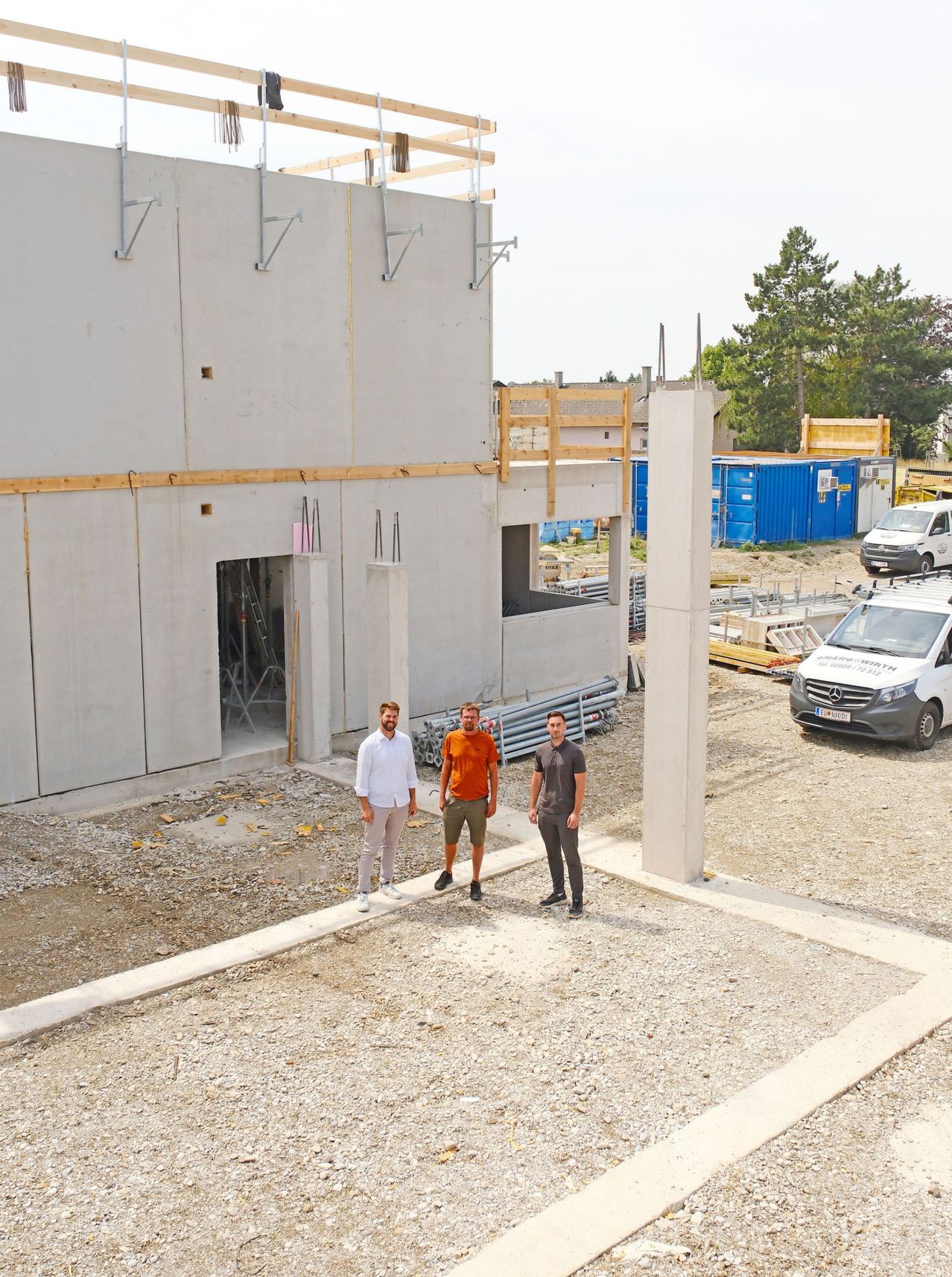 Drei Männer stehen vor einem teilweise errichteten Gebäude mit Betonwänden und Holzgerüst. Sie tragen Freizeitkleidung. Hinter ihnen ist eine Baustelle mit verschiedenen Materialien und Fahrzeugen zu sehen.