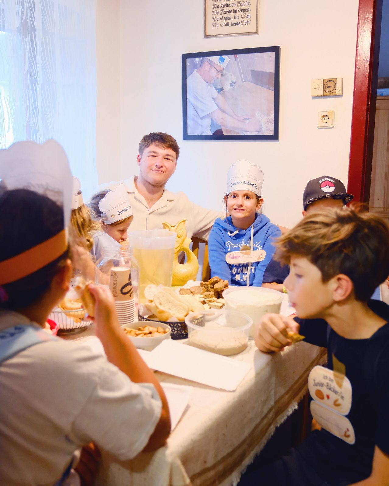 Eine Gruppe von Kindern und einem erwachsenen Mann sitzen um einen Tisch herum, tragen Kochmützen und essen. Der Tisch ist mit verschiedenen Speisen und Getränken gefüllt.