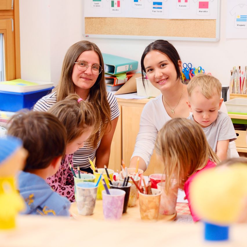 Zwei Frauen und mehrere Kinder sind in einem Klassenzimmer. Die Kinder sitzen um einen Tisch mit Malbechern und Pinseln. Eine Frau lächelt und schaut die Kinder an.