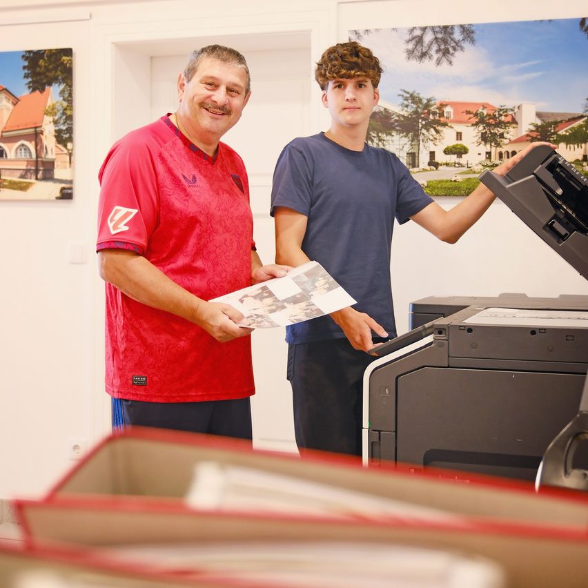 Ein Mann in einem roten Shirt und ein Junge in einem blauen Shirt stehen neben einem großen Drucker und halten ein Fotoalbum.