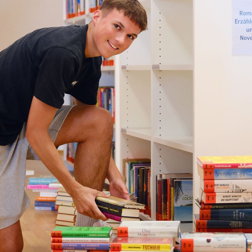 Ein junger Mann lächelt, während er Bücher von einem Regal in einer Bibliothek aufnimmt. Er trägt ein schwarzes T-Shirt und graue Shorts.