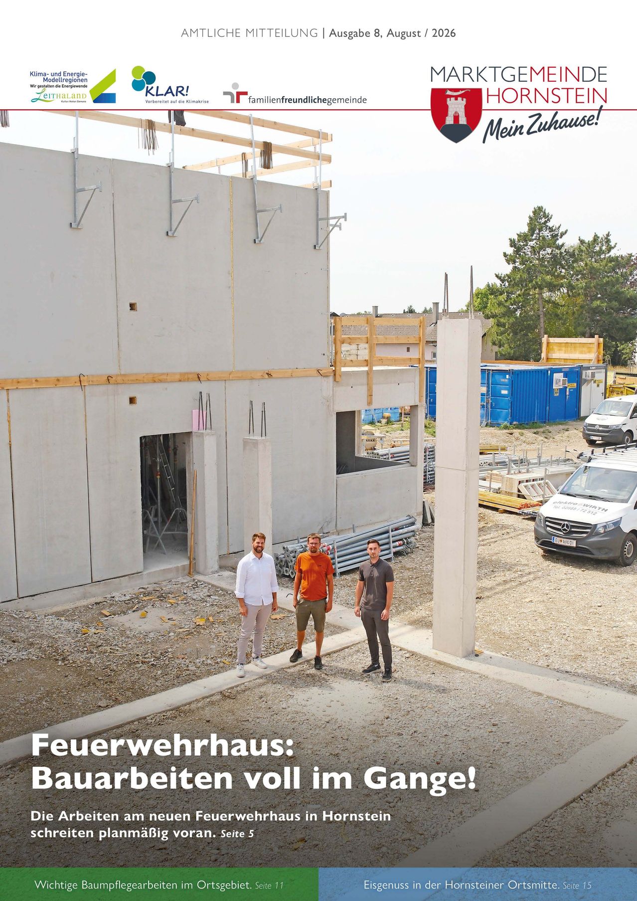 Drei Männer stehen vor einem teilweise errichteten Gebäude, möglicherweise einem Feuerwehrhaus, mit einem großen Pfeiler im Vordergrund und Baufahrzeugen im Hintergrund.
