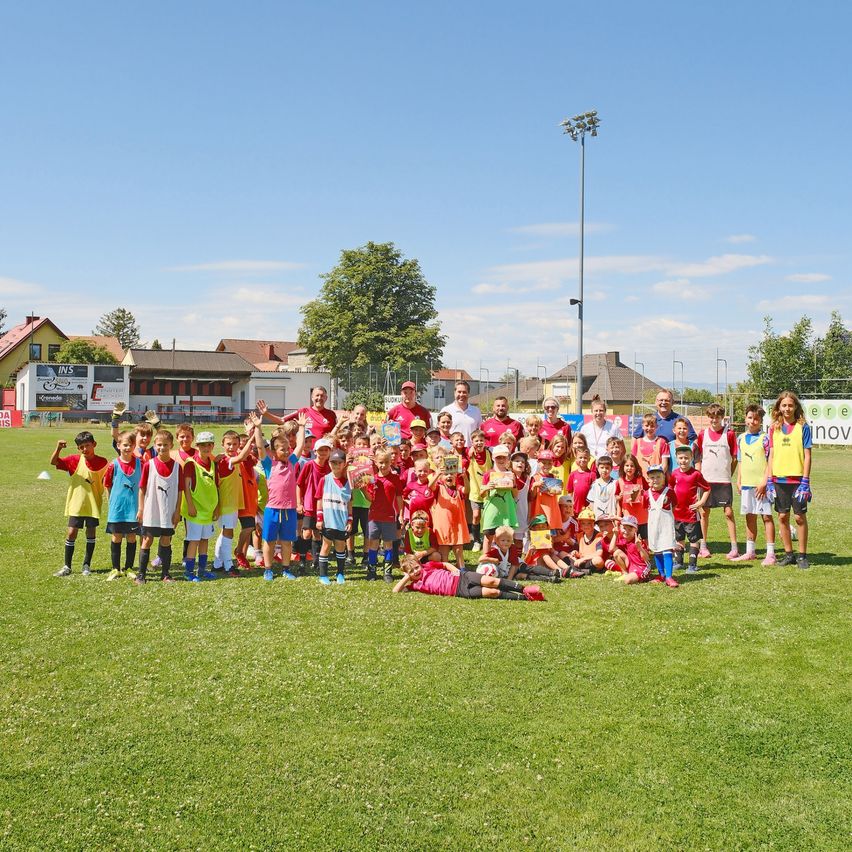 Eine große Gruppe von Kindern und Erwachsenen in Sportbekleidung posiert für ein Foto auf einem grünen Feld mit Bäumen und Gebäuden im Hintergrund.