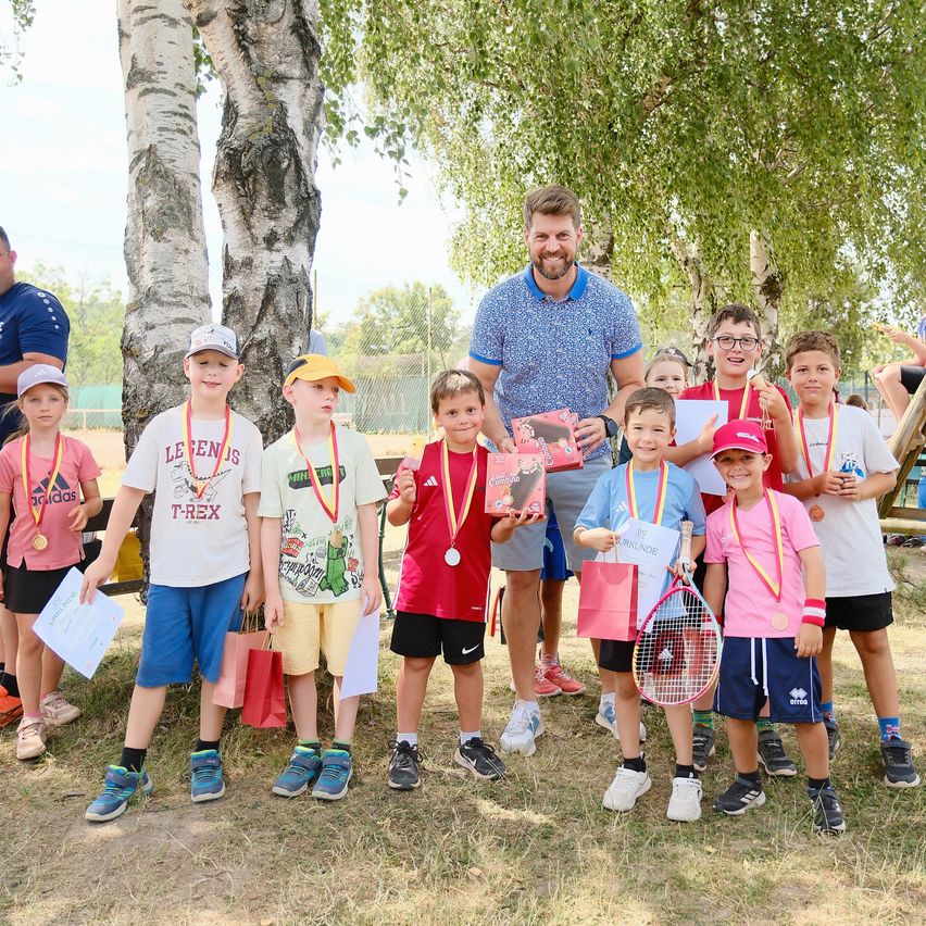 Eine Gruppe von Kindern mit Medaillen um den Hals steht neben einem Mann, der ein Buch und einen Tennisschläger hält. Sie alle lächeln und posieren für ein Foto.