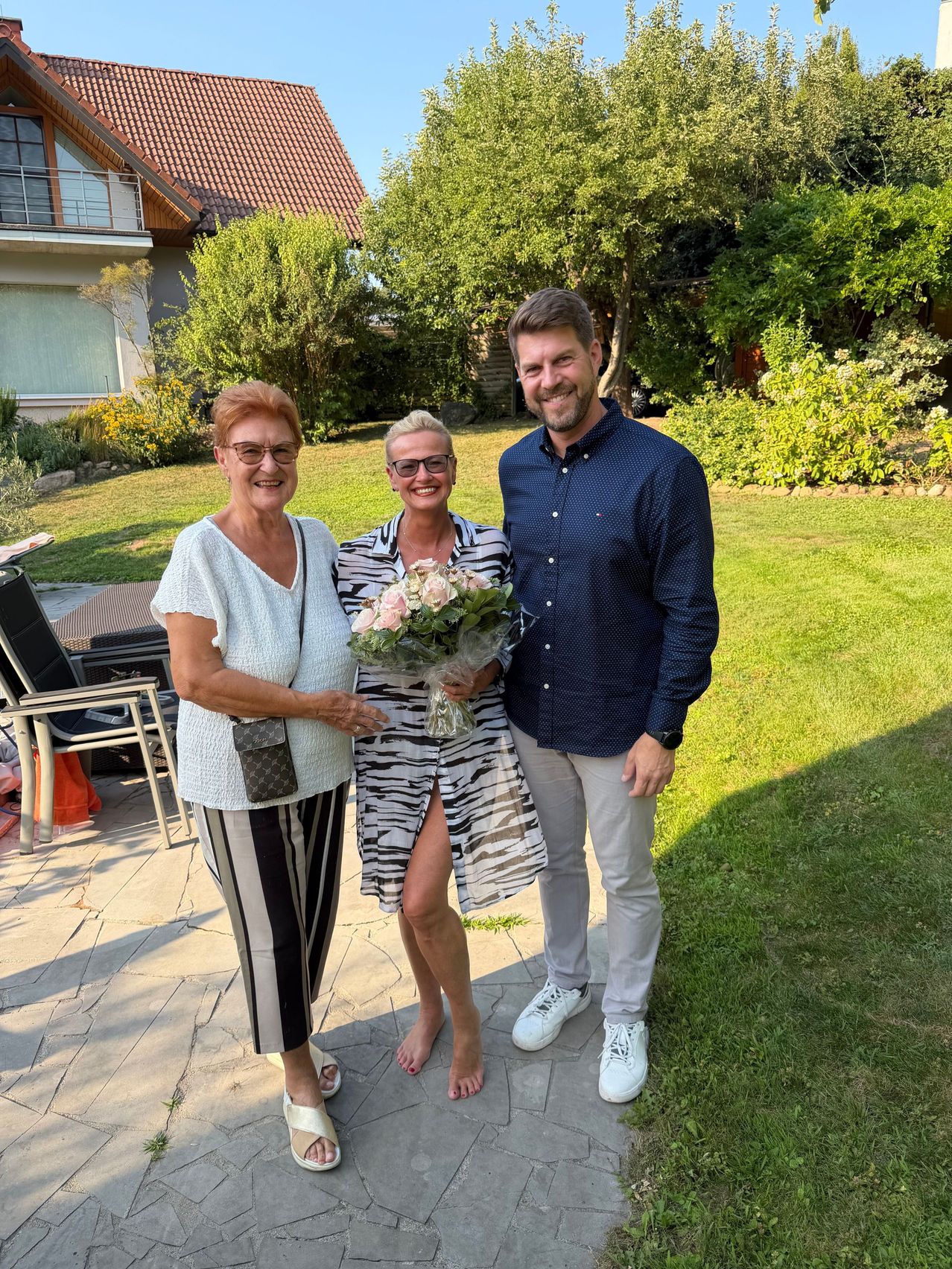 Three people stand in a garden, smiling. The woman on the left holds a handbag, the woman in the middle holds a bouquet of flowers, and the man on the right wears a watch.