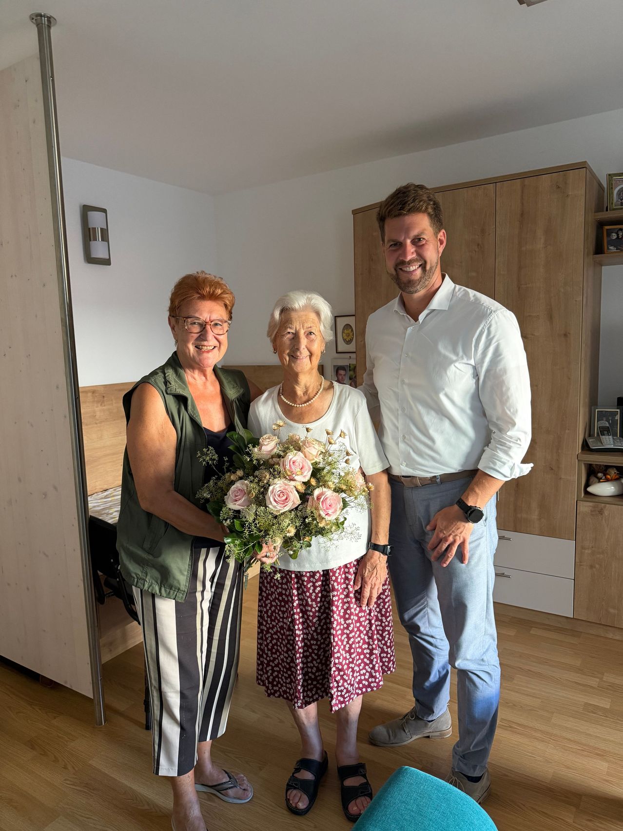Three people stand in a room, smiling. A woman holds a bouquet of flowers, and a man stands beside her. A woman on the left wears glasses and a green vest. Behind them is a wooden cabinet with picture frames.