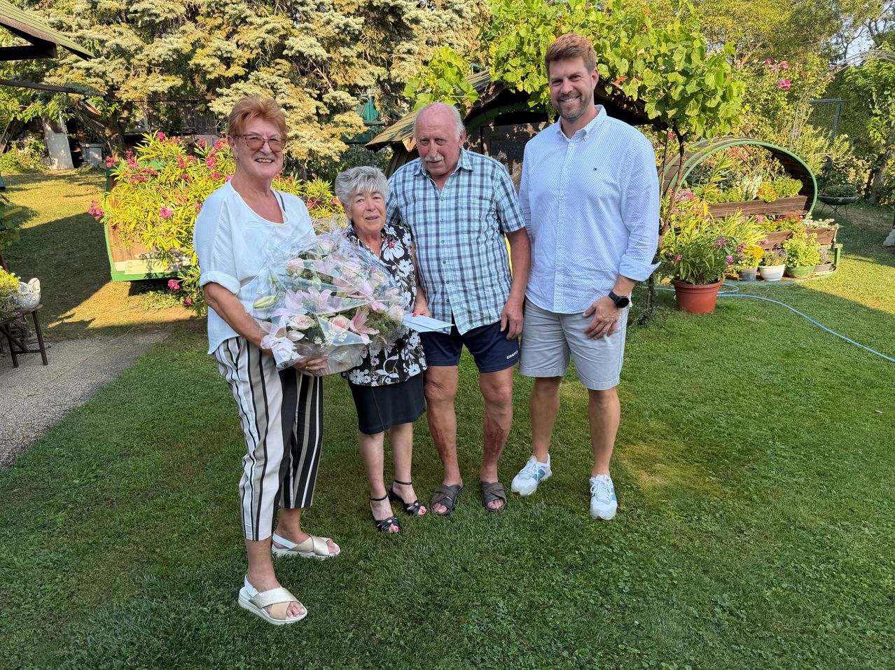 Four people stand in a garden, smiling. A woman holds a bouquet of flowers. Behind them are trees and plants.