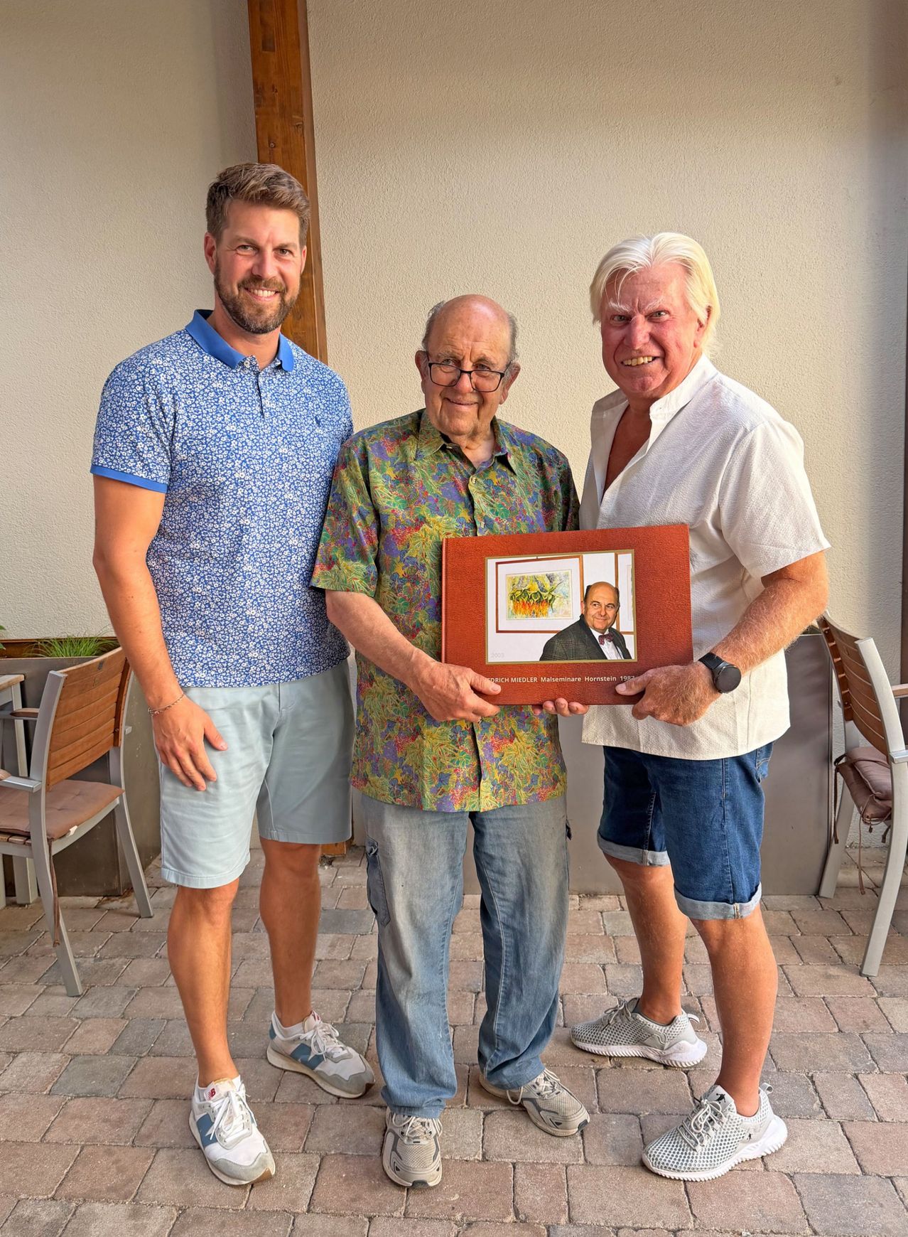 Three men stand together, one holding a framed portrait of a man in a suit and bow tie. The man in the middle wears glasses and a patterned shirt. The other two men wear casual attire.