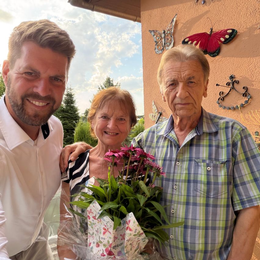 Three people stand together outside, smiling. A man on the left wears a white shirt, a woman in the middle holds flowers, and an older man on the right wears a plaid shirt. Behind them is a wall with butterfly decorations.