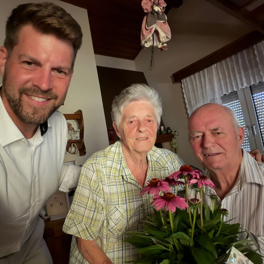 Three people smiling in a room with flowers in the foreground, a window with blinds, and a shelf with items in the background.