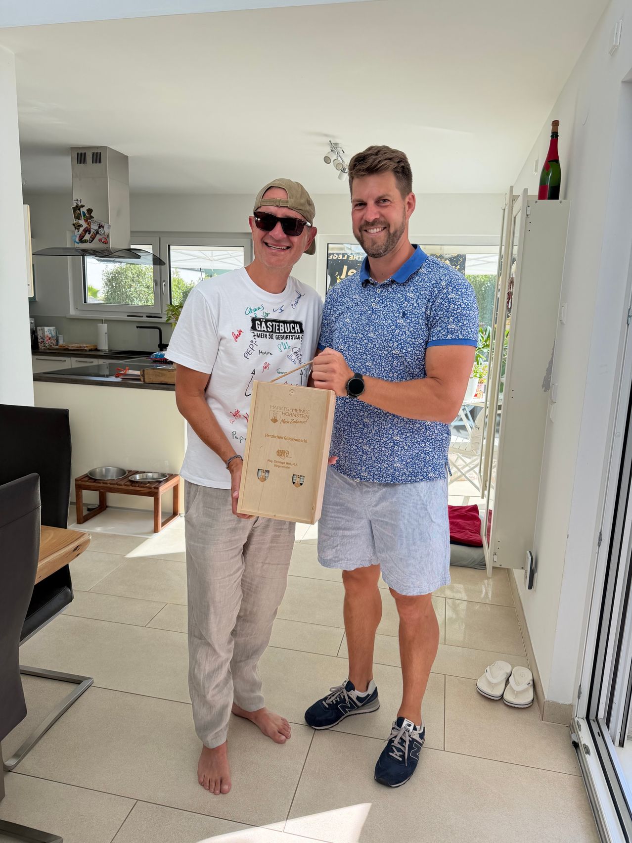 Two men are standing in a room, one holding a certificate in a brown paper bag. The man on the left is wearing a white shirt with a design, while the man on the right is wearing a blue polo shirt.