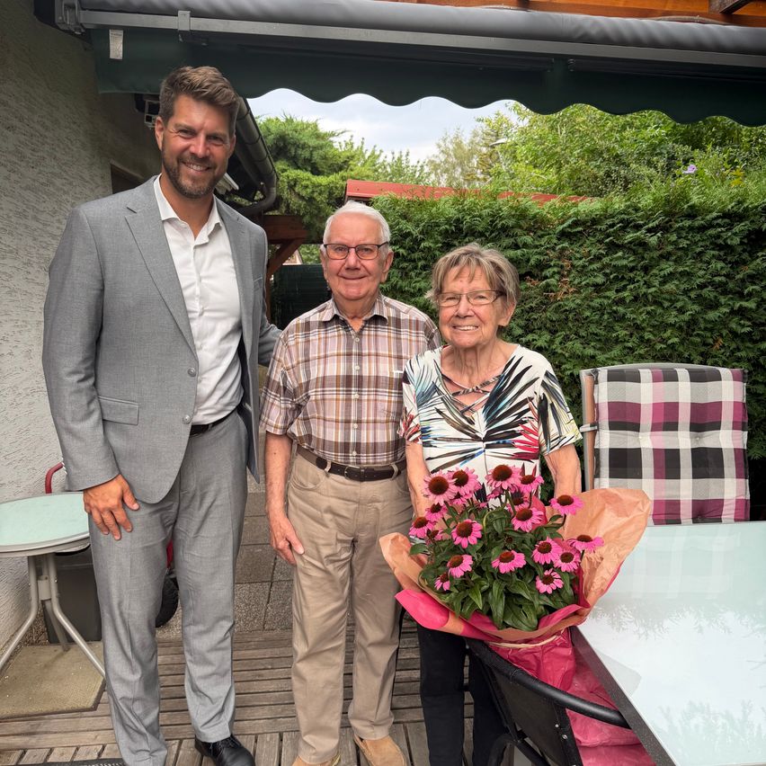 Drei Menschen stehen auf einer Terrasse und lächeln. Ein Mann in einem grauen Anzug, ein Mann in einem karierten Hemd und eine Frau mit Blumen. Dahinter steht ein Busch und ein Haus.