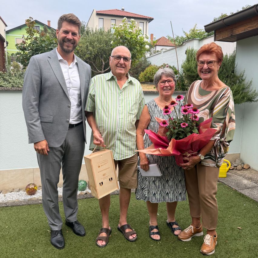 Vier Personen stehen auf einem grünen Rasen und lächeln für ein Foto. Ein Mann im Anzug hält eine Schachtel, während ein Mann in einem gestreiften Hemd einen Blumenstrauß hält. Zwei Frauen stehen neben ihnen, eine hält ein Papier.